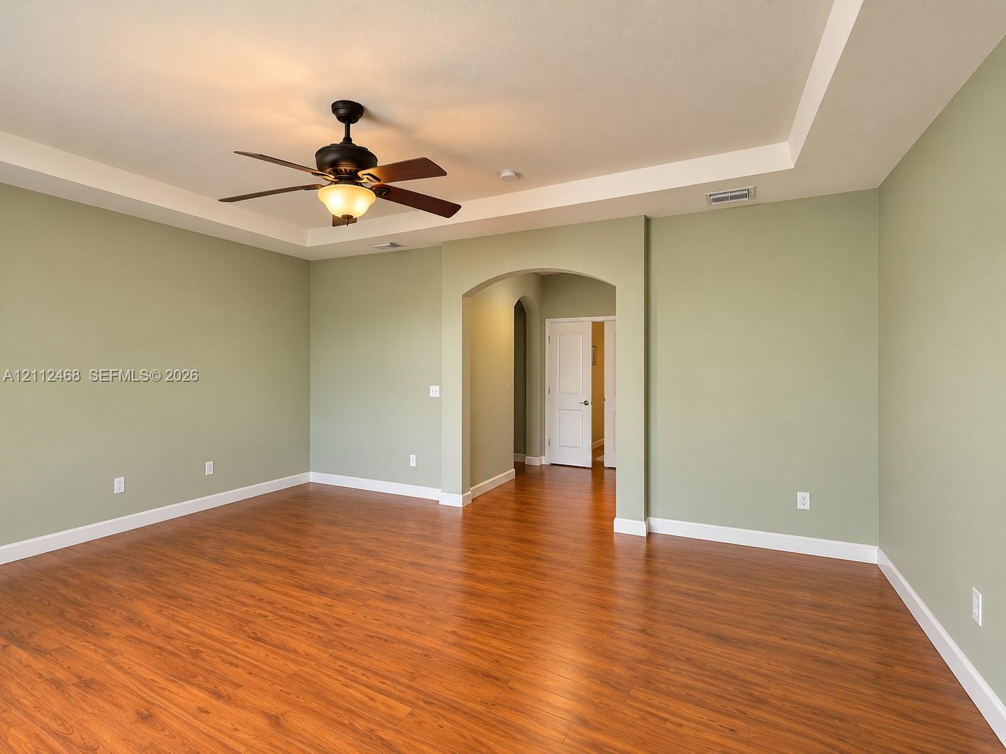 Room featuring wood-finish flooring, a tray ceiling, an arched entryway, and an attached ceiling fan with a light fixture
