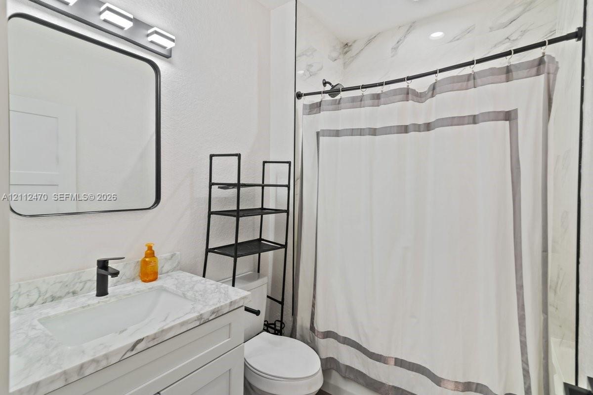 Bathroom vanity with stone-finish countertop, undermount sink, matte black faucet, rectangular mirror with a black frame, and shower surround with stone-finish wall tile