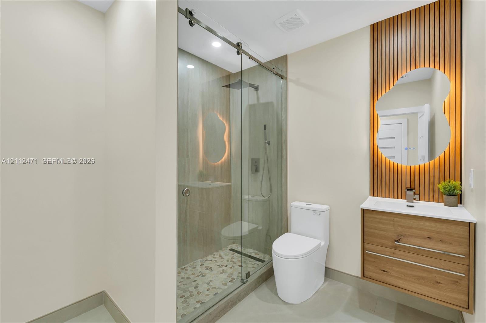 Glass-enclosed shower with a pebble-stone floor, wood-finish floating vanity, vertical wood-slat accent wall, and irregular-shaped backlit mirror