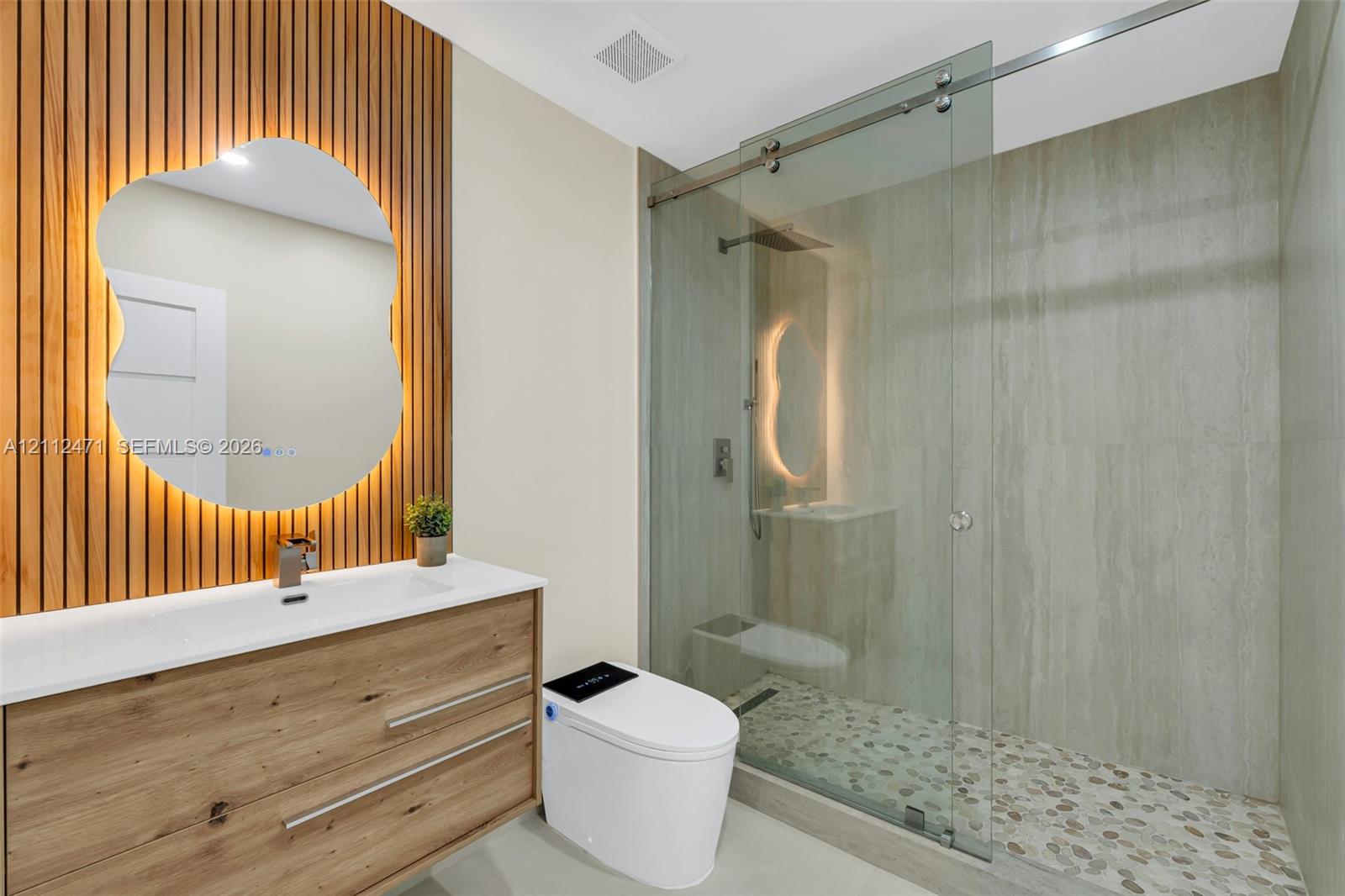 Wood-slat accent wall featuring a backlit, organic-shaped mirror, wood-finish vanity with an integrated basin, and a walk-in shower with a glass enclosure and pebble-stone flooring