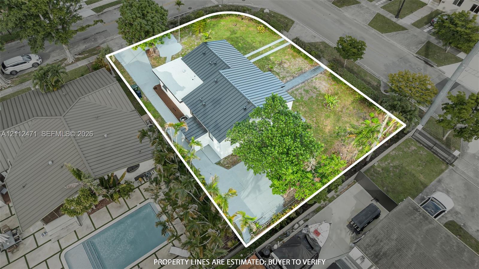 Aerial view featuring a metal roof, concrete driveway, and expansive grass lot with mature trees