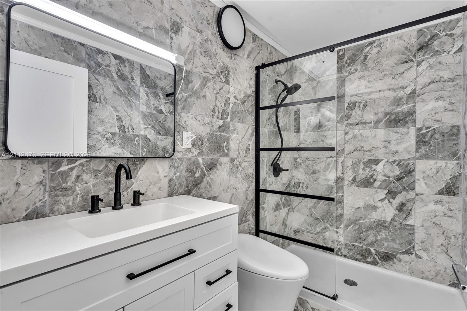 Bathroom featuring full-height stone-finish tile surround, white vanity with quartz countertop, matte black fixtures, framed vanity mirror, and glass shower enclosure with black grid detailing