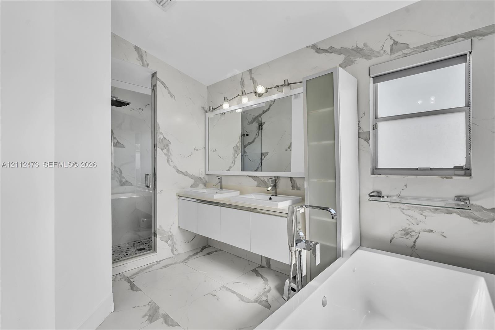 Bathroom featuring floor-to-ceiling large-format stone-patterned wall tile, white floating double vanity with integrated sinks, glass-enclosed walk-in shower, soaking tub, and built-in vertical storage cabinet