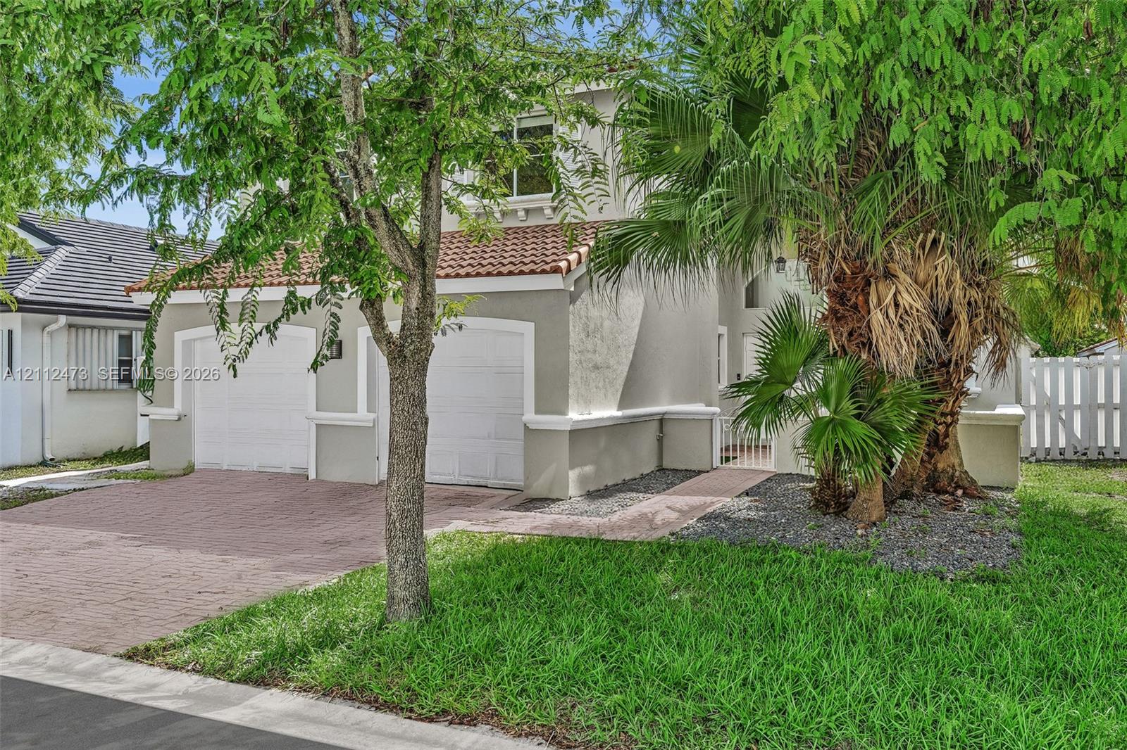 Stucco exterior featuring a two-car garage, red clay tile roofing, decorative trim detailing, and a paved brick driveway