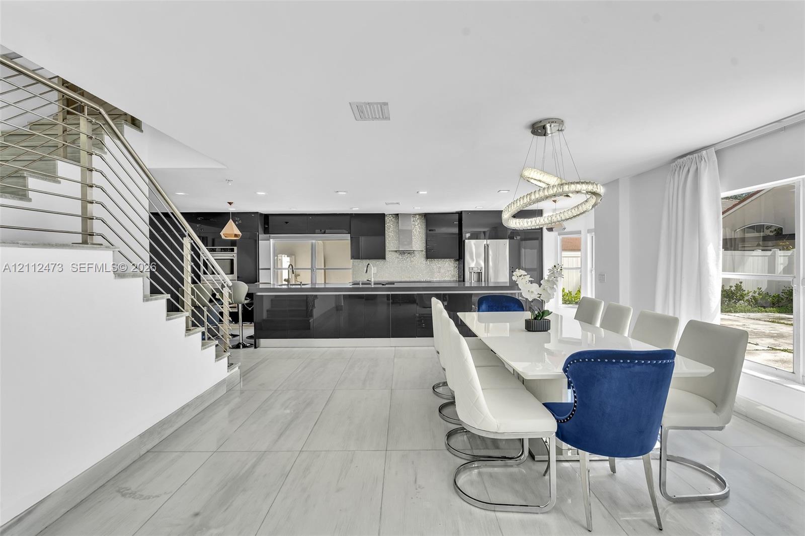 Open-concept interior featuring large-format tile flooring, high-gloss cabinetry, stainless steel range hood, stainless steel horizontal railing staircase, and a modern circular LED chandelier