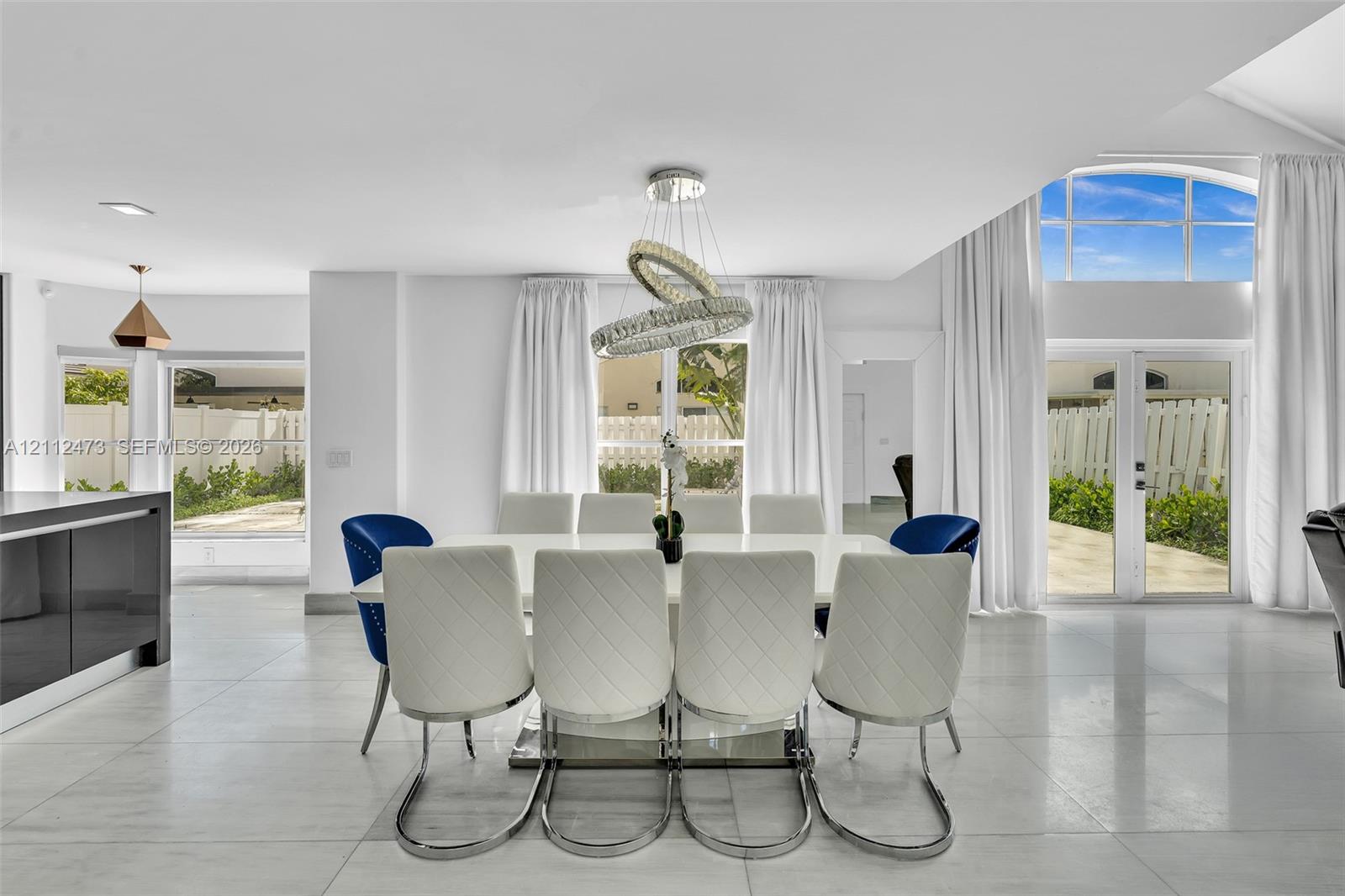 Open-concept interior featuring large-format tile flooring, a contemporary multi-ring crystal chandelier, vaulted ceilings, and dual sets of glass-paned doors with arched transom windows