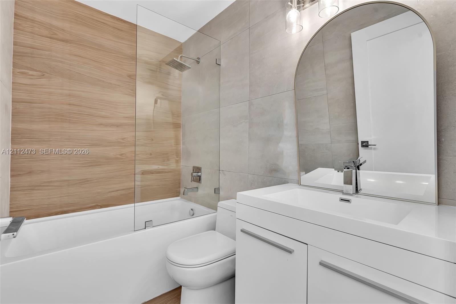 Bathroom featuring a floating white vanity with a rectangular sink, arch-top mirror, wood-finish wall tile, frameless glass shower panel, and large-format neutral wall tile