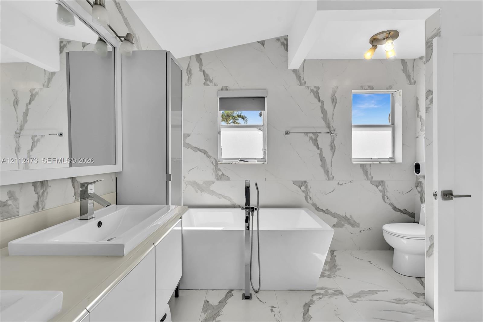 Bathroom featuring floor-to-ceiling large-format tile with decorative veining, freestanding soaking tub with floor-mounted faucet, rectangular vessel sink atop a modern vanity, and dual wall-mounted light fixtures