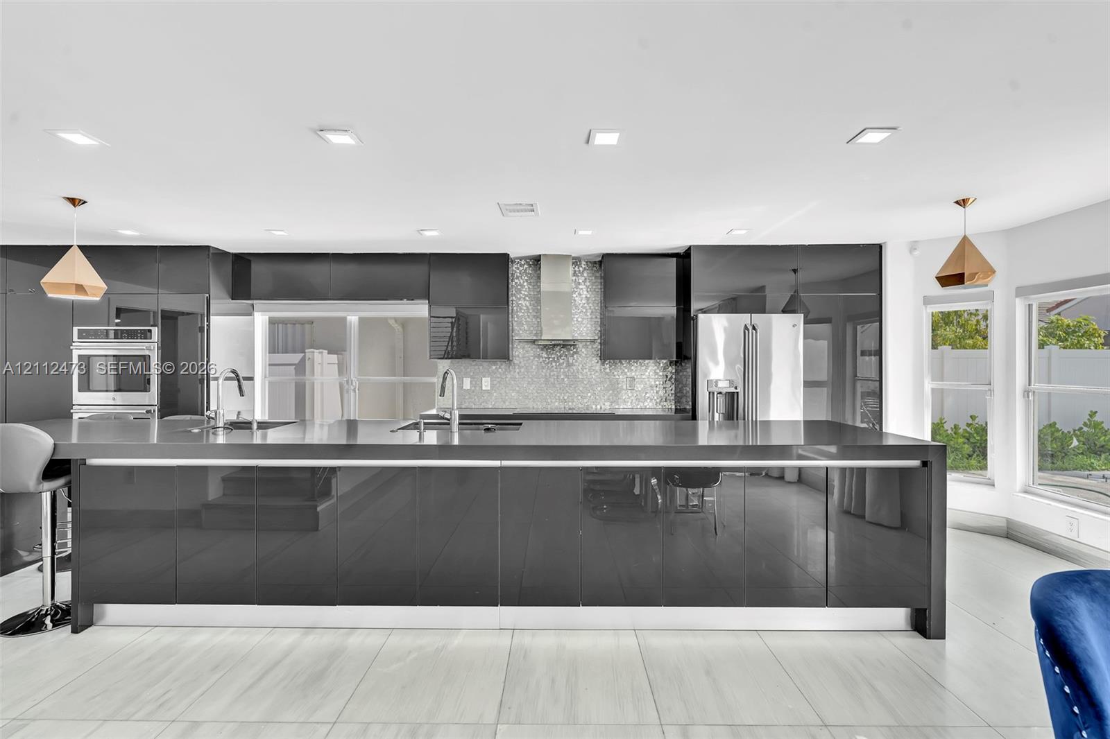 Modern kitchen featuring high-gloss cabinetry, a spacious island with a waterfall edge countertop, mosaic tile backsplash, stainless steel appliances, and recessed lighting