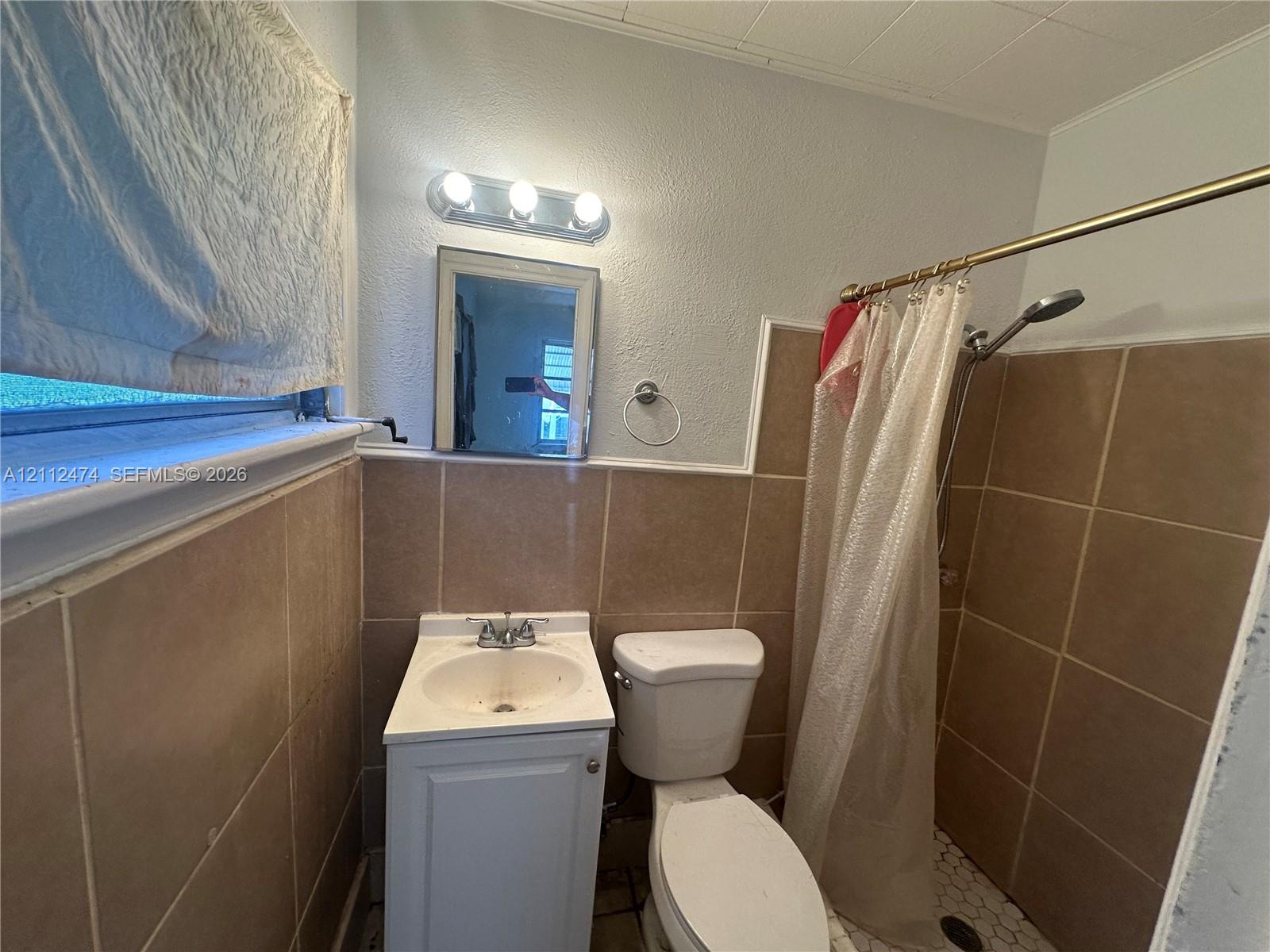 Bathroom featuring neutral-tone square wall tile, pedestal-style vanity with integrated sink, three-light wall-mounted fixture, and a walk-in shower with hexagonal floor tile Bathroom featuring neutral-tone square wall tile, pedestal-style vanity with integrated sink, three-light wall-mounted fixture, and a walk-in shower with hexagonal floor tile