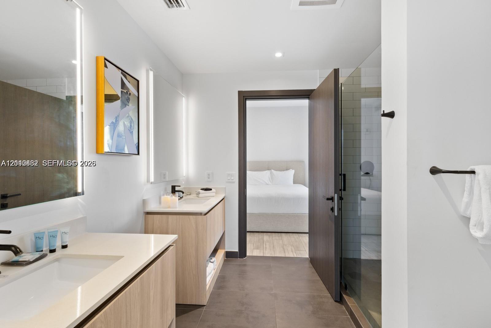 Dual vanity with light wood-finish cabinetry, solid surface countertops, integrated LED-lit mirrors, and glass-enclosed shower with subway tile