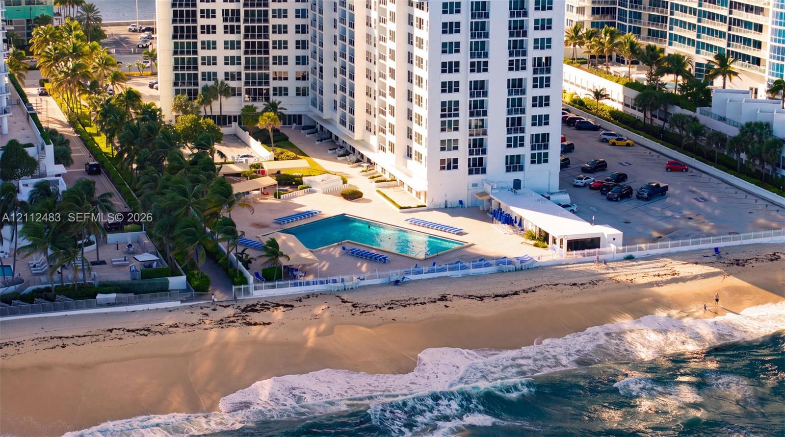 High-rise residential building featuring an L-shaped swimming pool, paved sundeck, direct beach access, and surface parking