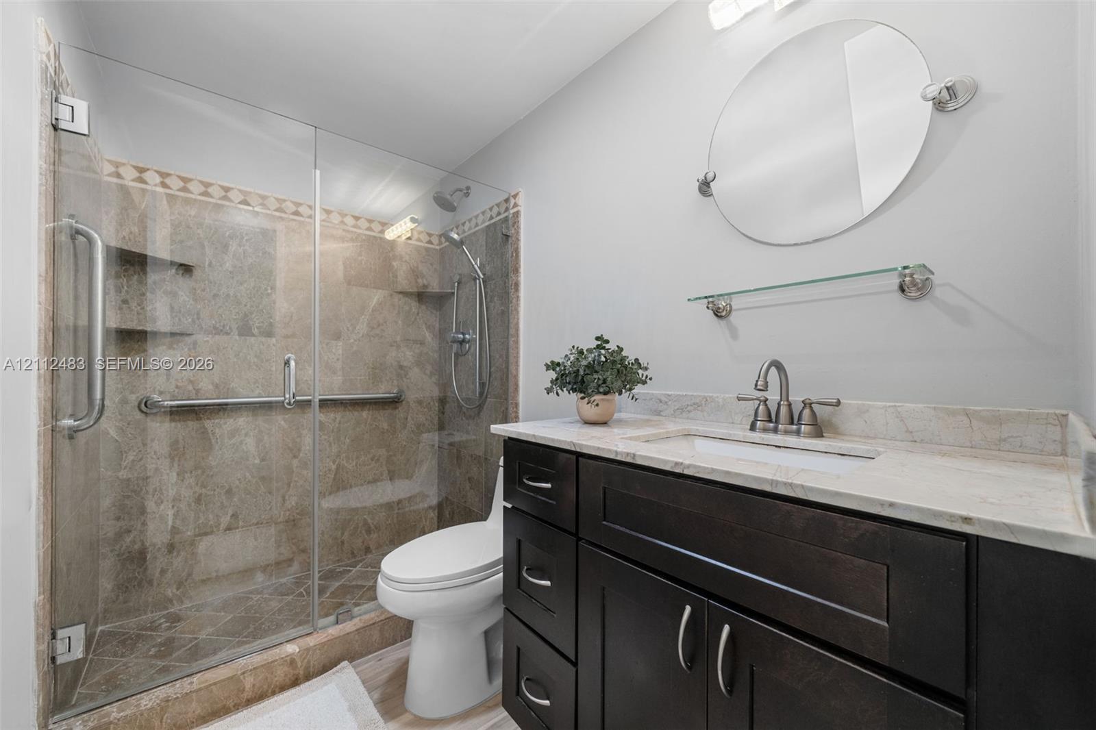 Dark wood vanity with a stone countertop, frameless glass shower enclosure, stone-tiled shower surround with built-in shelving, adjustable handheld shower head, and an oval wall-mounted mirror