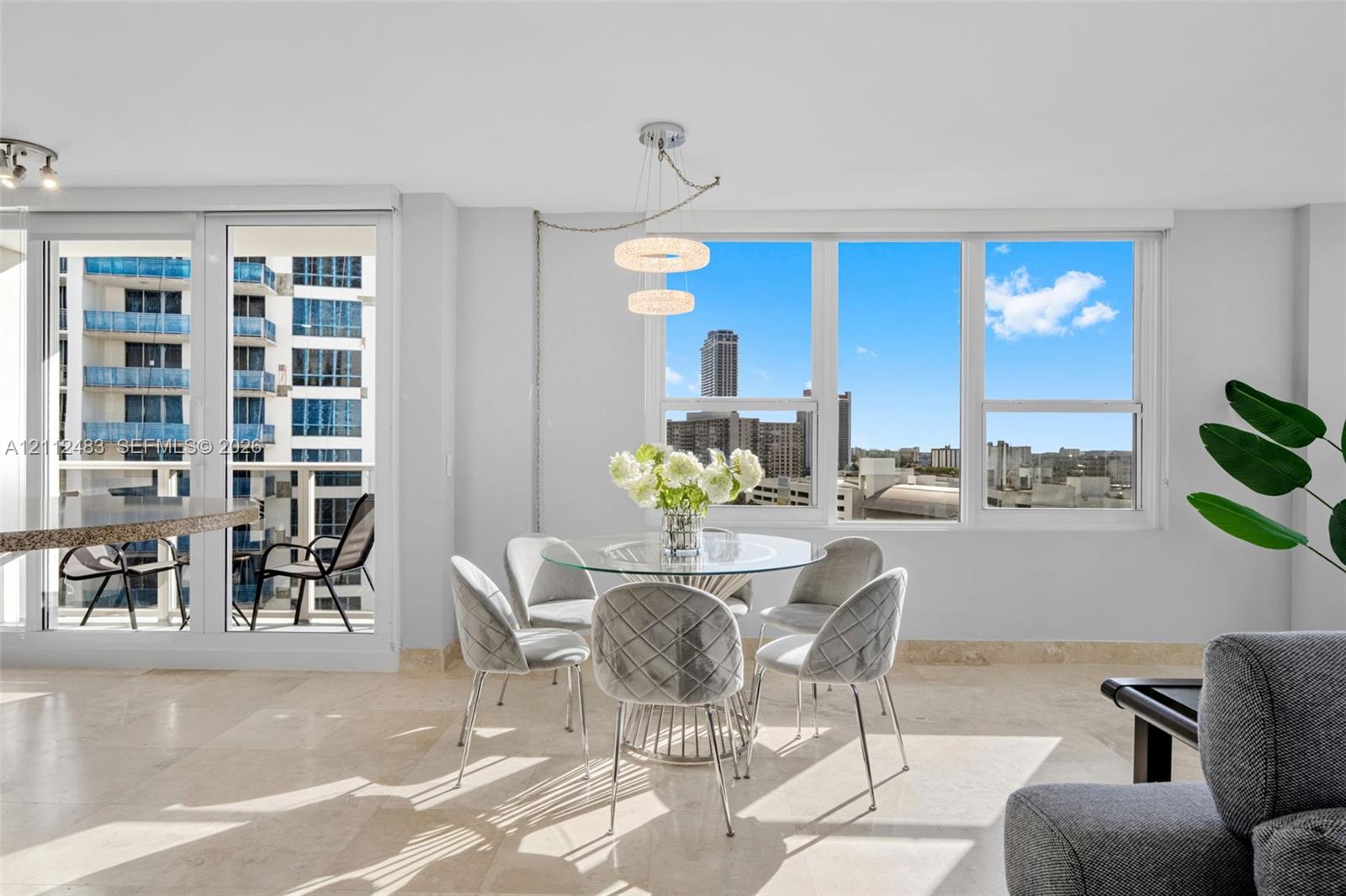 Light-filled interior featuring floor-to-ceiling windows, natural stone flooring, a multi-tier contemporary chandelier, and a glass-paneled door opening to a private balcony