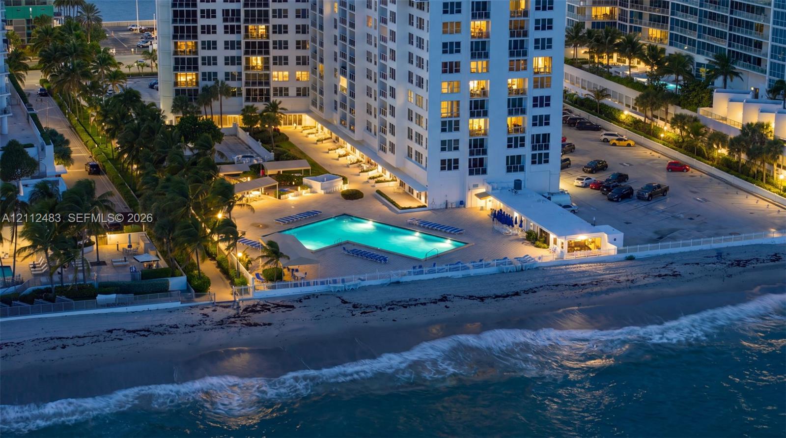 High-rise building featuring a swimming pool, paved sundeck, landscaped grounds with mature palm trees, and direct oceanfront positioning