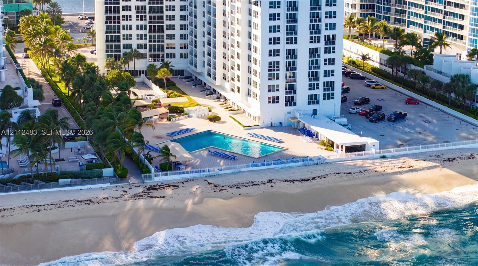High-rise residential building featuring direct beach access, an L-shaped swimming pool, paved surface parking, and mature palm landscaping