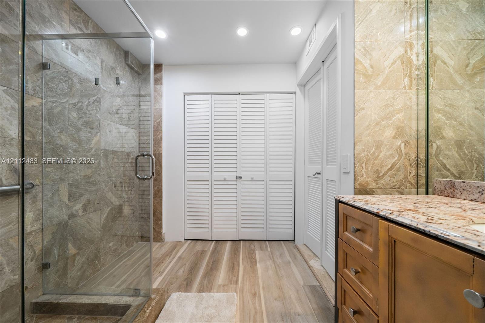 Walk-in shower with stone tile surround and glass enclosure, wood-finish flooring, stone-topped vanity with built-in cabinetry, and white louvered closet doors