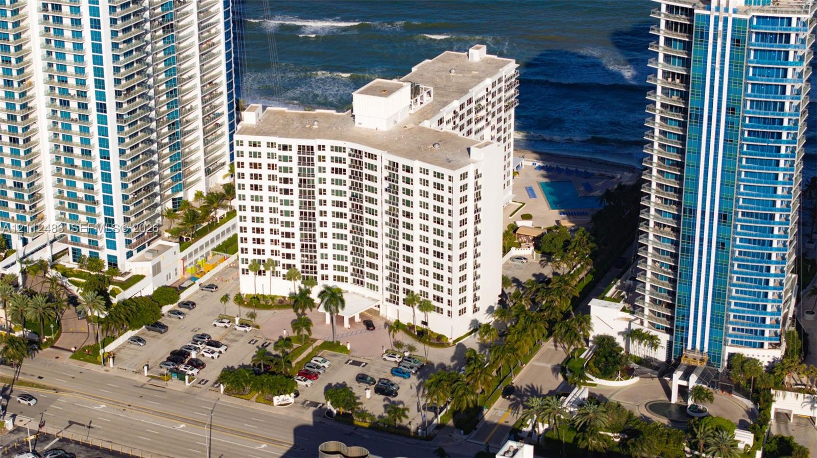 Aerial perspective of high-rise building featuring proximity to the coastline, paved surface parking, mature palm trees, and an outdoor swimming pool