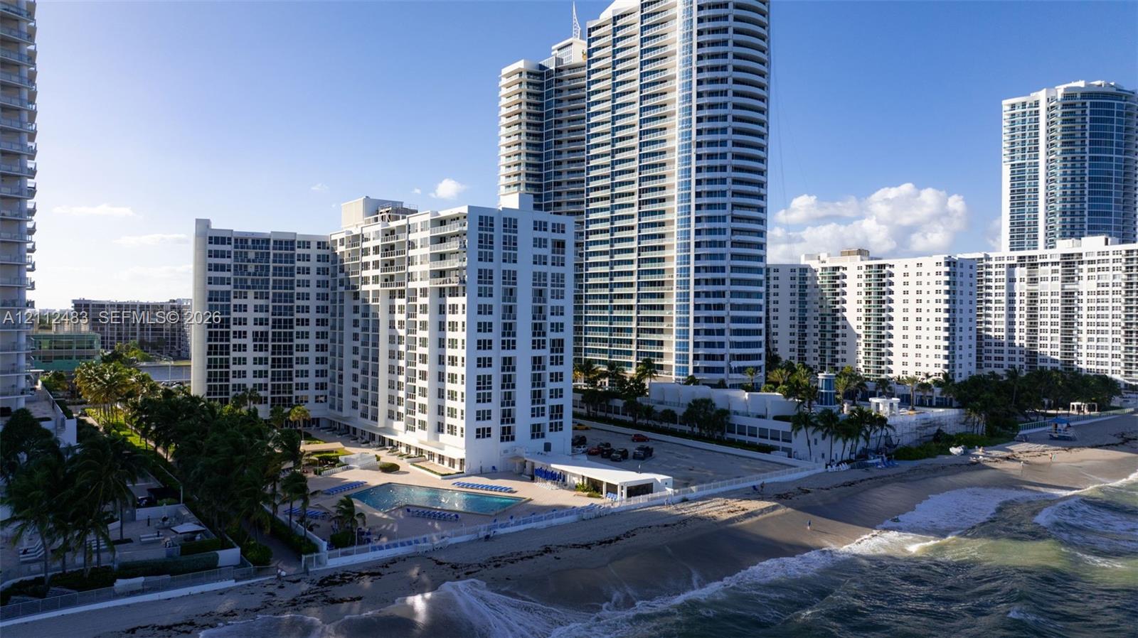 High-rise residential building featuring expansive beachfront frontage, an outdoor swimming pool, paved surface parking, and mature palm trees