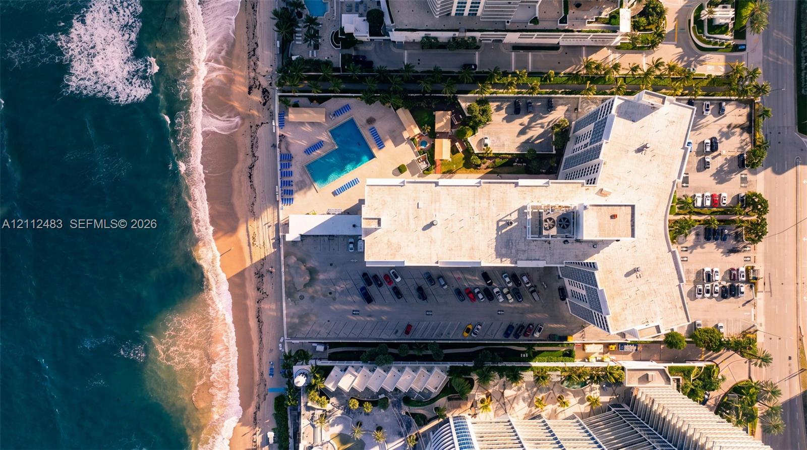 Aerial perspective featuring direct oceanfront access, a coastal pool, surface parking, and manicured grounds