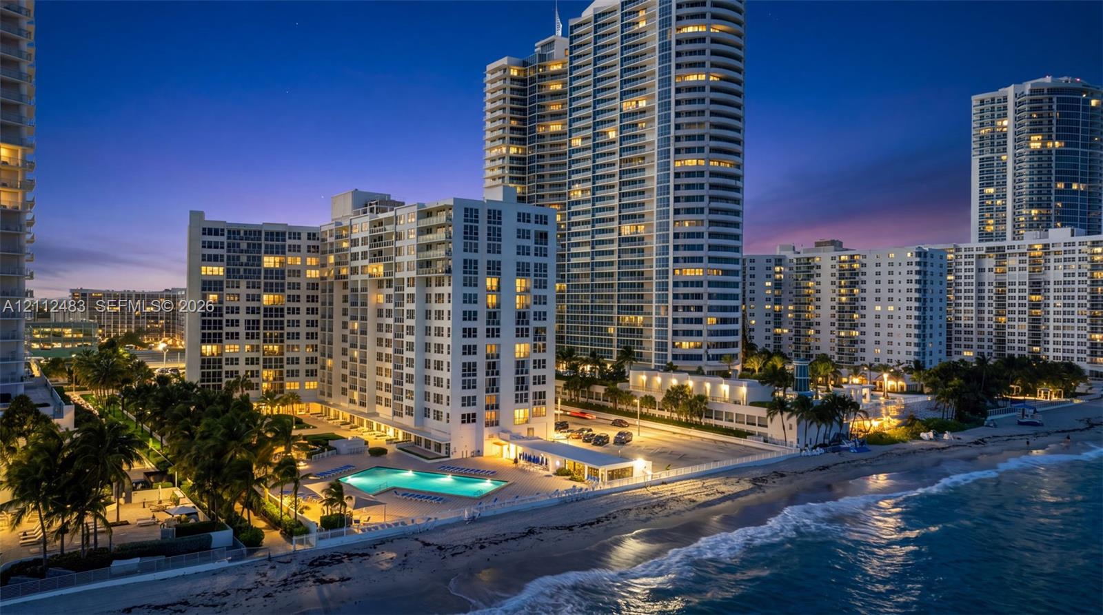 High-rise residential complex featuring direct beach access, an outdoor swimming pool, integrated parking area, and palm tree landscaping