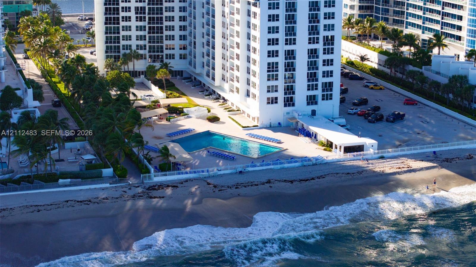 High-rise residential building featuring a waterfront swimming pool, paved sundeck, landscaped grounds with palm trees, and direct oceanfront access