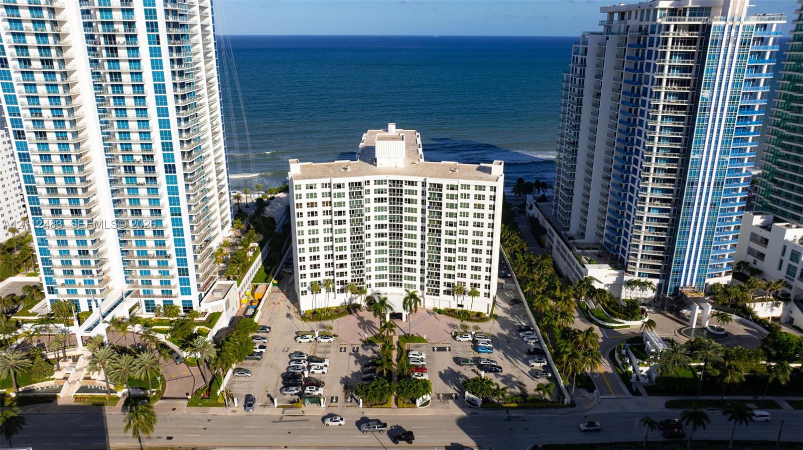 Mid-rise residential building featuring a concrete exterior, expansive glass windows, ground-level parking, and direct oceanfront positioning
