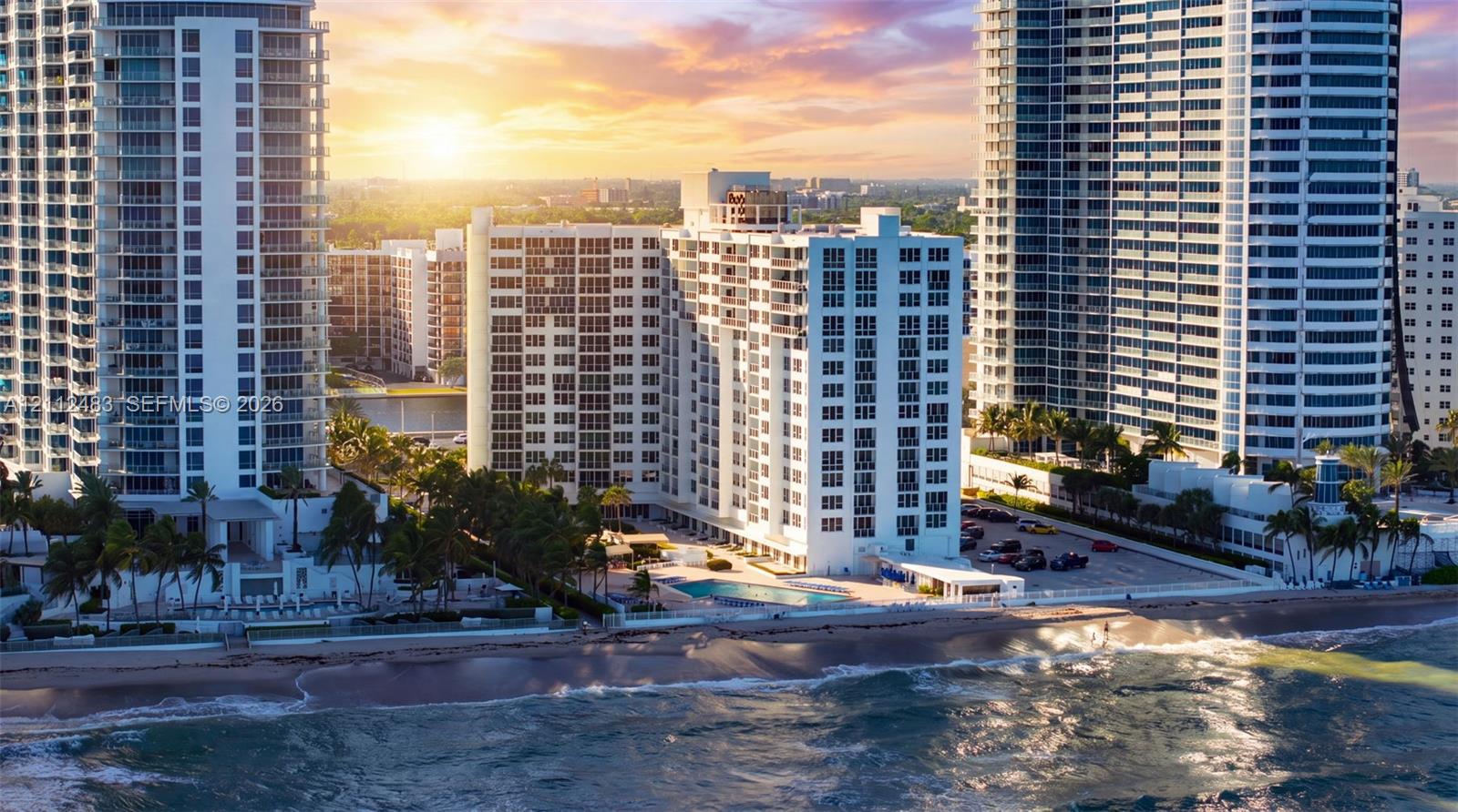 High-rise building exterior featuring direct oceanfront positioning, ground-level pool, paved surface parking, and mature palm landscaping
