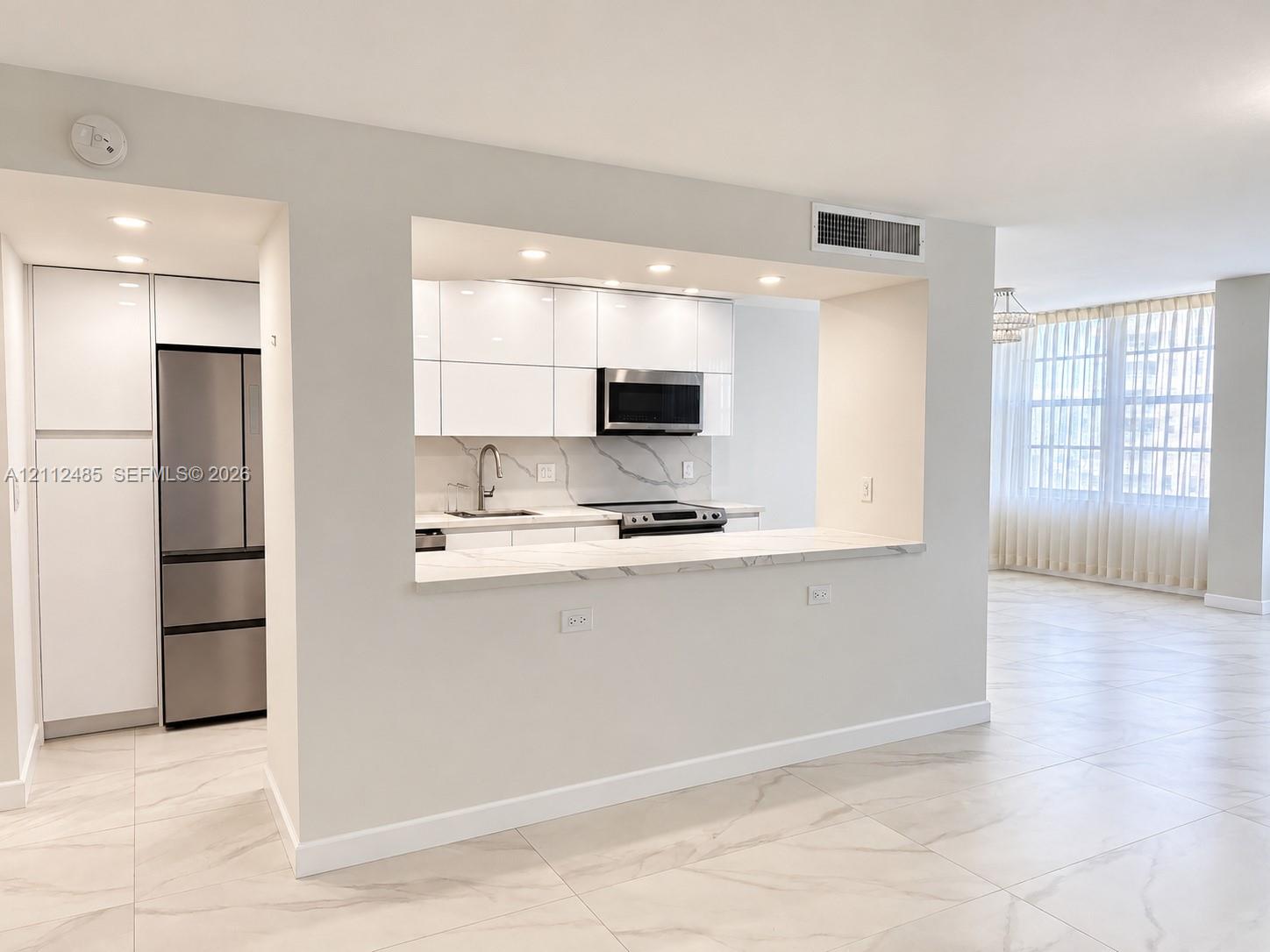 Kitchen featuring high-gloss white cabinetry, stone-finish countertops with waterfall edge, stainless steel refrigerator, built-in microwave, and large-format tile flooring Kitchen featuring high-gloss white cabinetry, stone-finish countertops with waterfall edge, stainless steel refrigerator, built-in microwave, and large-format tile flooring
