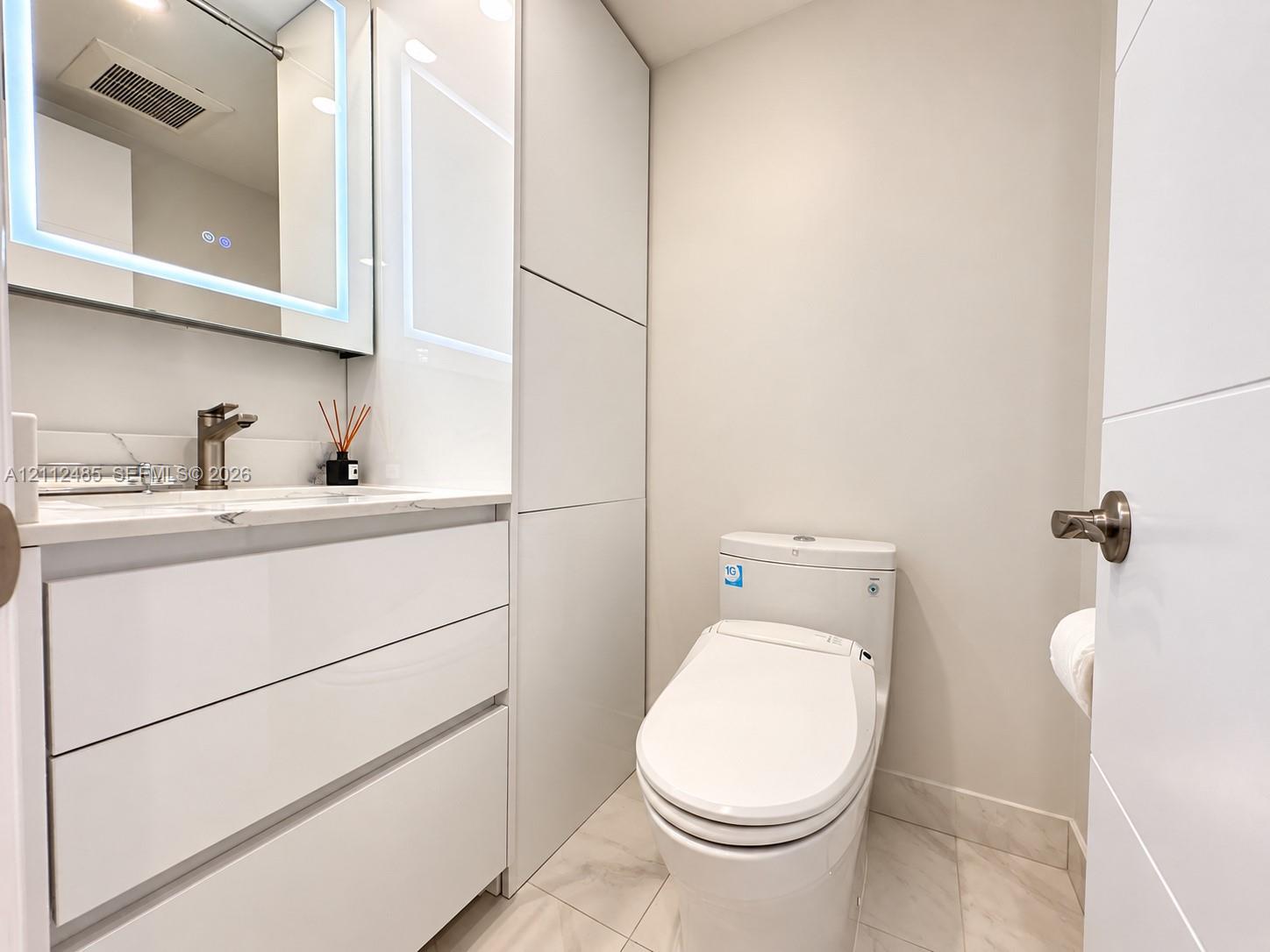 Modern vanity with integrated storage, stone-surfaced countertop, LED-backlit mirror, and neutral tile flooring