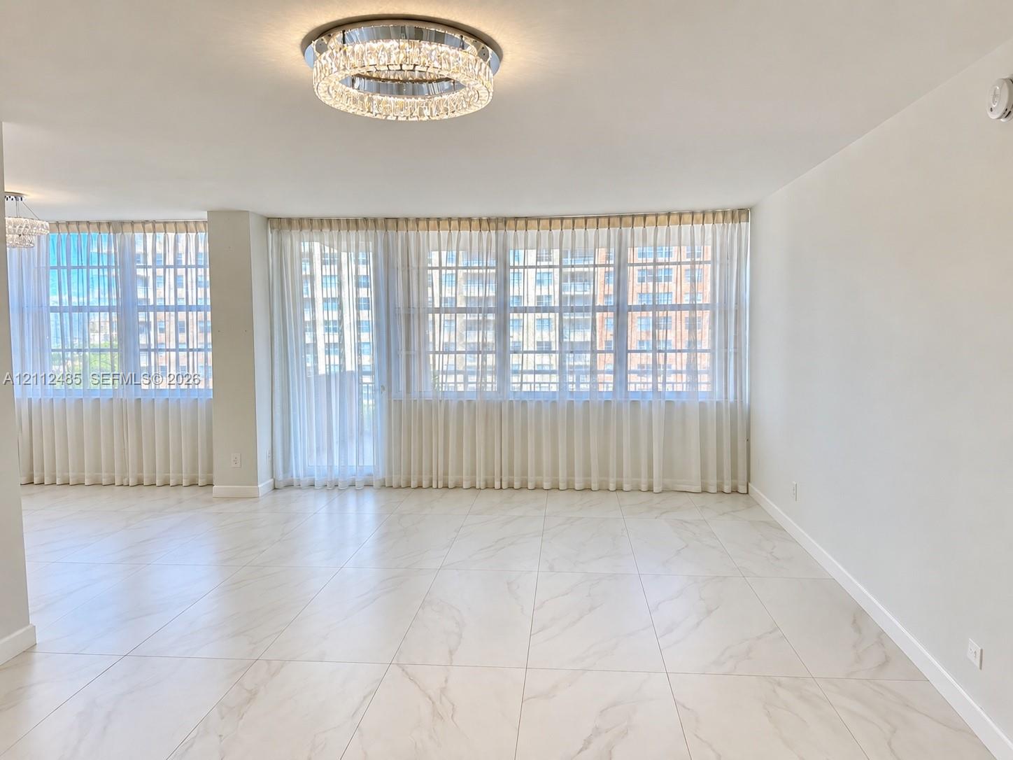 Open-concept interior featuring large-format tile flooring with marble-style veining, floor-to-ceiling windows, and a tiered crystal flush-mount ceiling fixture Open-concept interior featuring large-format tile flooring with marble-style veining, floor-to-ceiling windows, and a tiered crystal flush-mount ceiling fixture