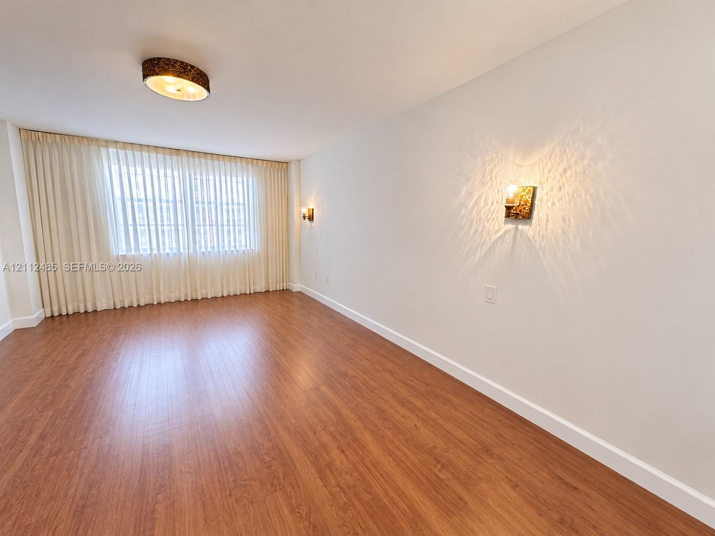 Neutral interior featuring wood-finish flooring, a drum-style ceiling fixture, wall-mounted decorative sconces, and full-length window treatments