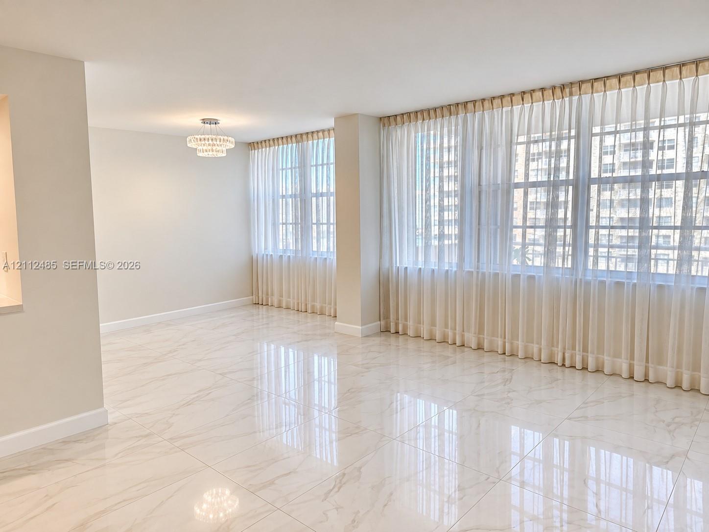 Large-format neutral stone-finish tile flooring, expansive windows with wall-to-wall drapery, and a tiered crystal-style flush-mount ceiling fixture Large-format neutral stone-finish tile flooring, expansive windows with wall-to-wall drapery, and a tiered crystal-style flush-mount ceiling fixture