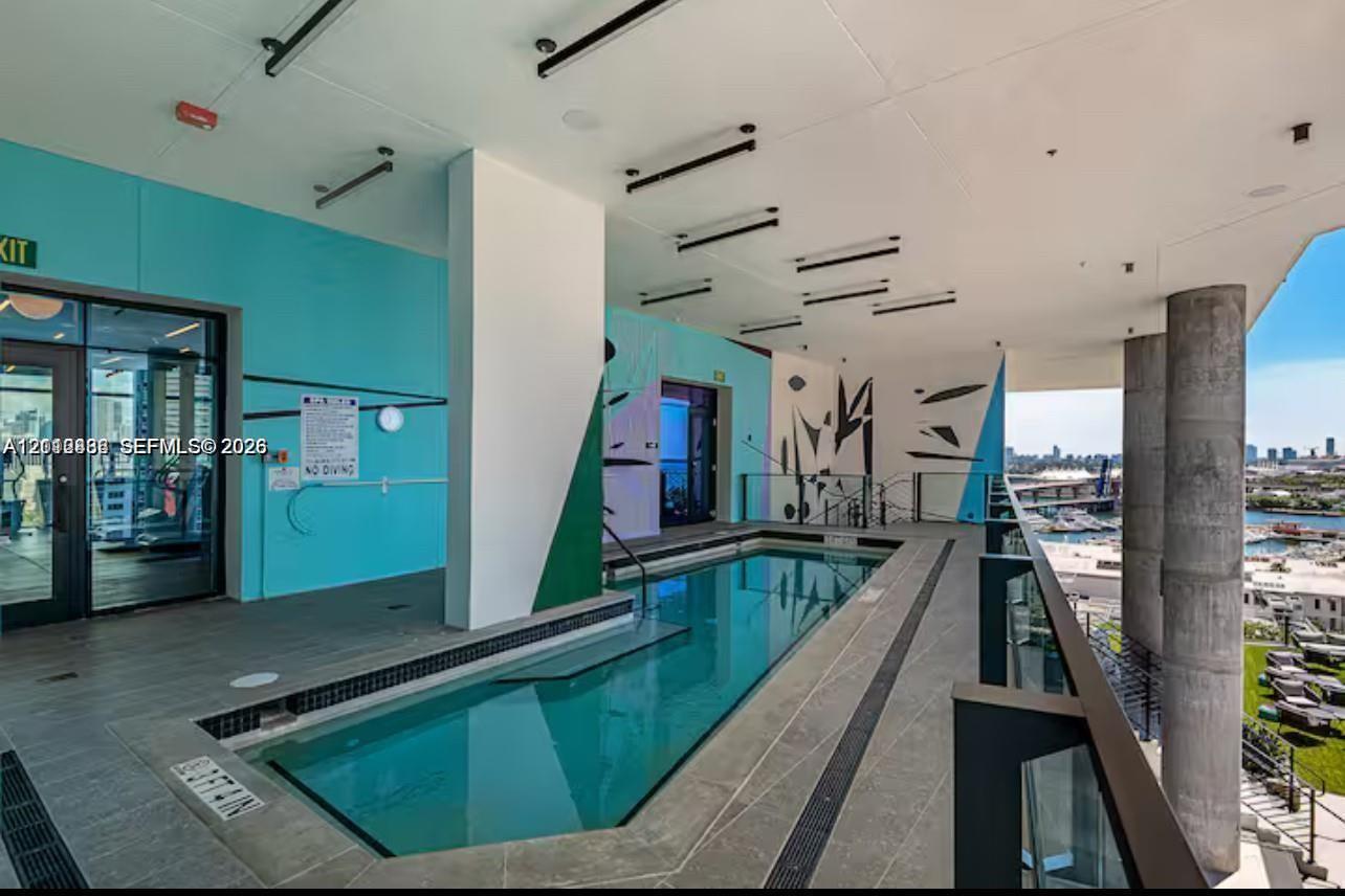 Covered outdoor pool featuring grey stone-finish tile decking, structural concrete support columns, a glass railing system, and linear recessed lighting