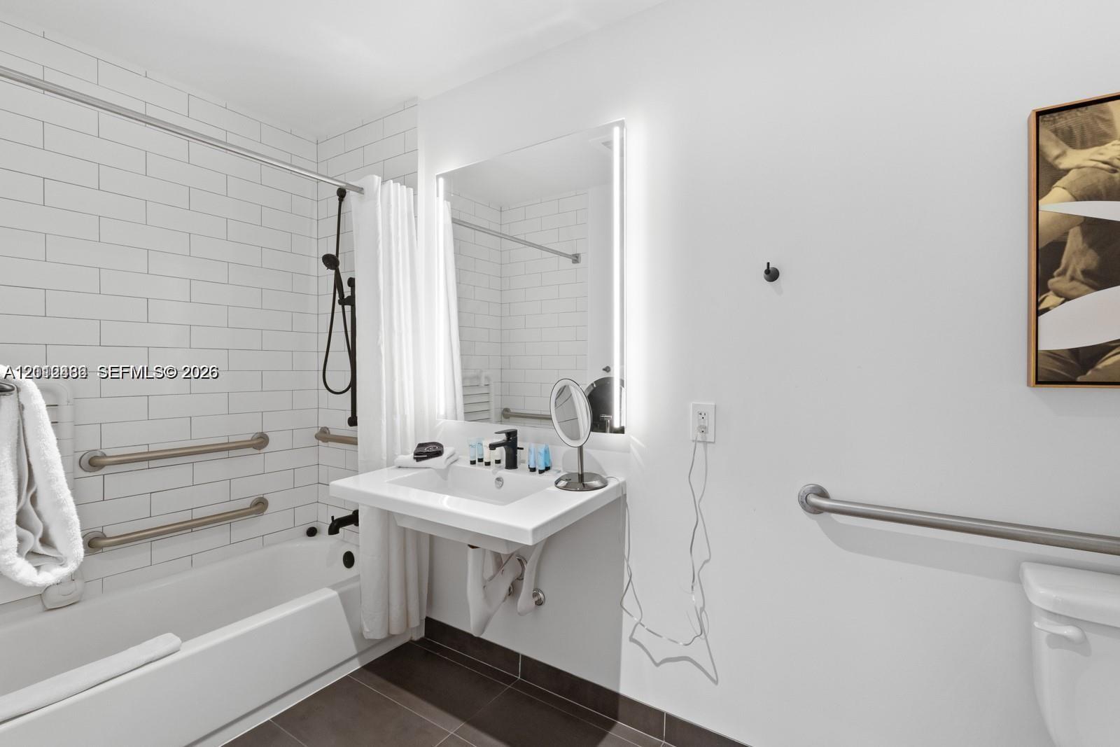 Wall-mounted floating vanity with integrated sink, rectangular illuminated mirror, white subway-tiled shower surround, matte black plumbing fixtures, and installed safety grab bars Wall-mounted floating vanity with integrated sink, rectangular illuminated mirror, white subway-tiled shower surround, matte black plumbing fixtures, and installed safety grab bars