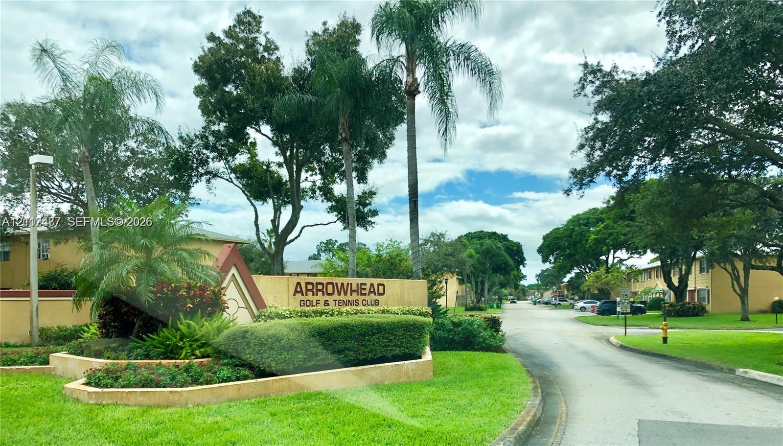 Community entrance featuring stucco monument signage, manicured landscaping with tropical palm trees, paved roadway, and mature canopy trees Community entrance featuring stucco monument signage, manicured landscaping with tropical palm trees, paved roadway, and mature canopy trees