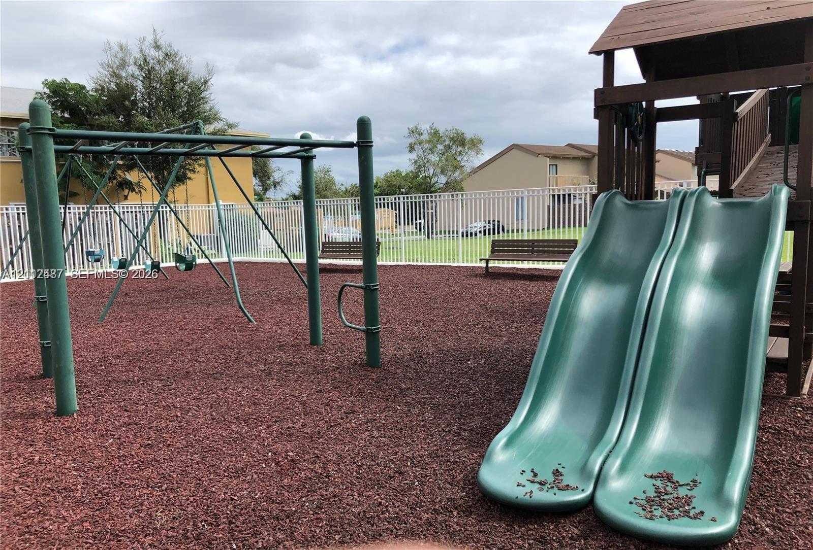 Outdoor recreation area featuring a multi-station jungle gym, dual slides, a swing set, and metal bench seating situated on a rubberized surface