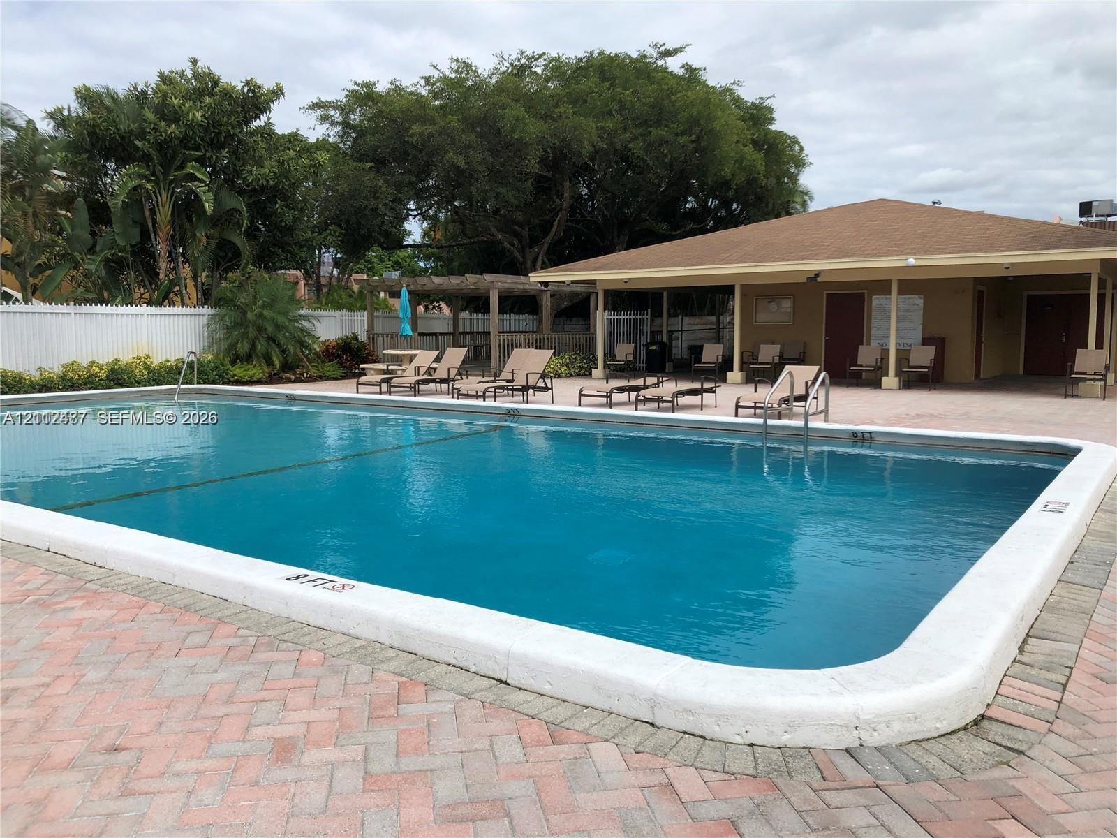 In-ground swimming pool featuring a concrete deck, herringbone-pattern brick paver surrounds, a wooden pergola, and a covered clubhouse structure