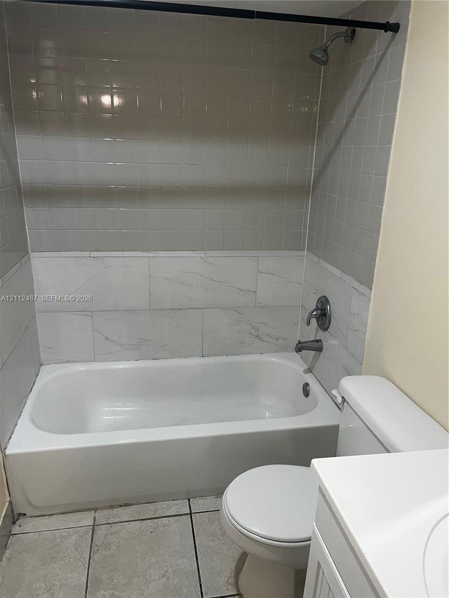 Full bathroom featuring a white porcelain tub with a two-tone tiled surround, integrated shower fixture, tile flooring, and a white vanity Full bathroom featuring a white porcelain tub with a two-tone tiled surround, integrated shower fixture, tile flooring, and a white vanity