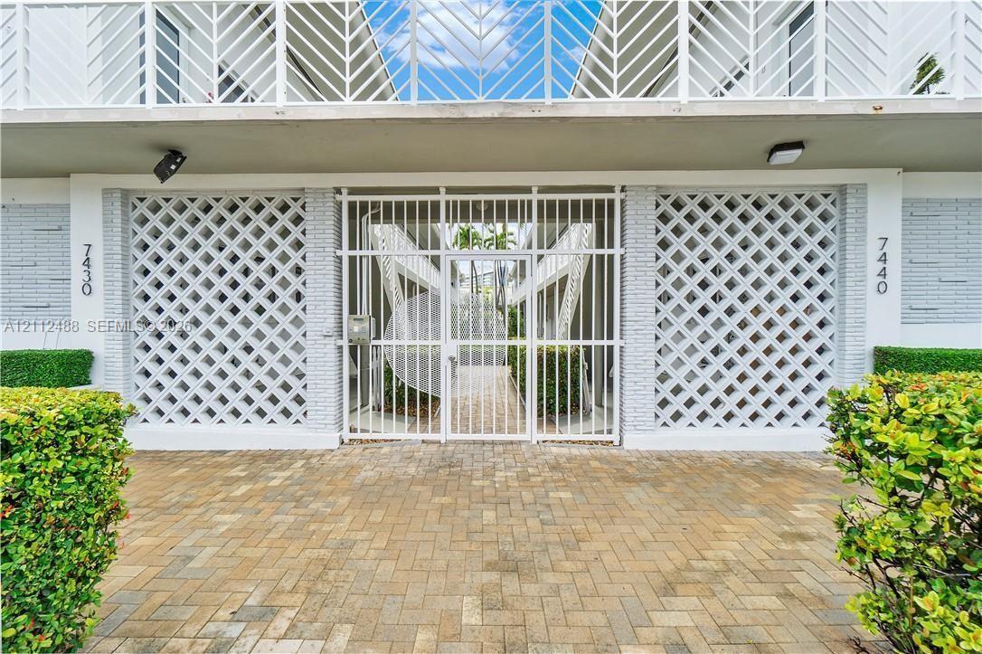 Exterior facade featuring decorative white breeze block accents, a secured metal gate entrance, a brick-paver entryway, and white chevron-patterned balcony railings