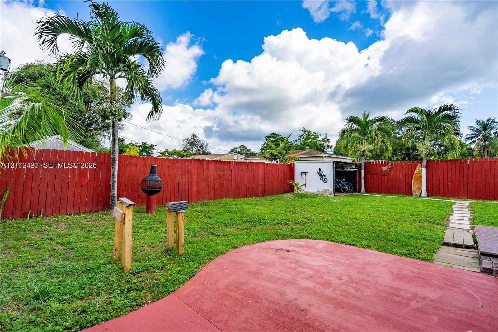 Backyard featuring a red-painted concrete patio, mature palm trees, a grass lawn, a wood-slat privacy fence, and a detached storage shed