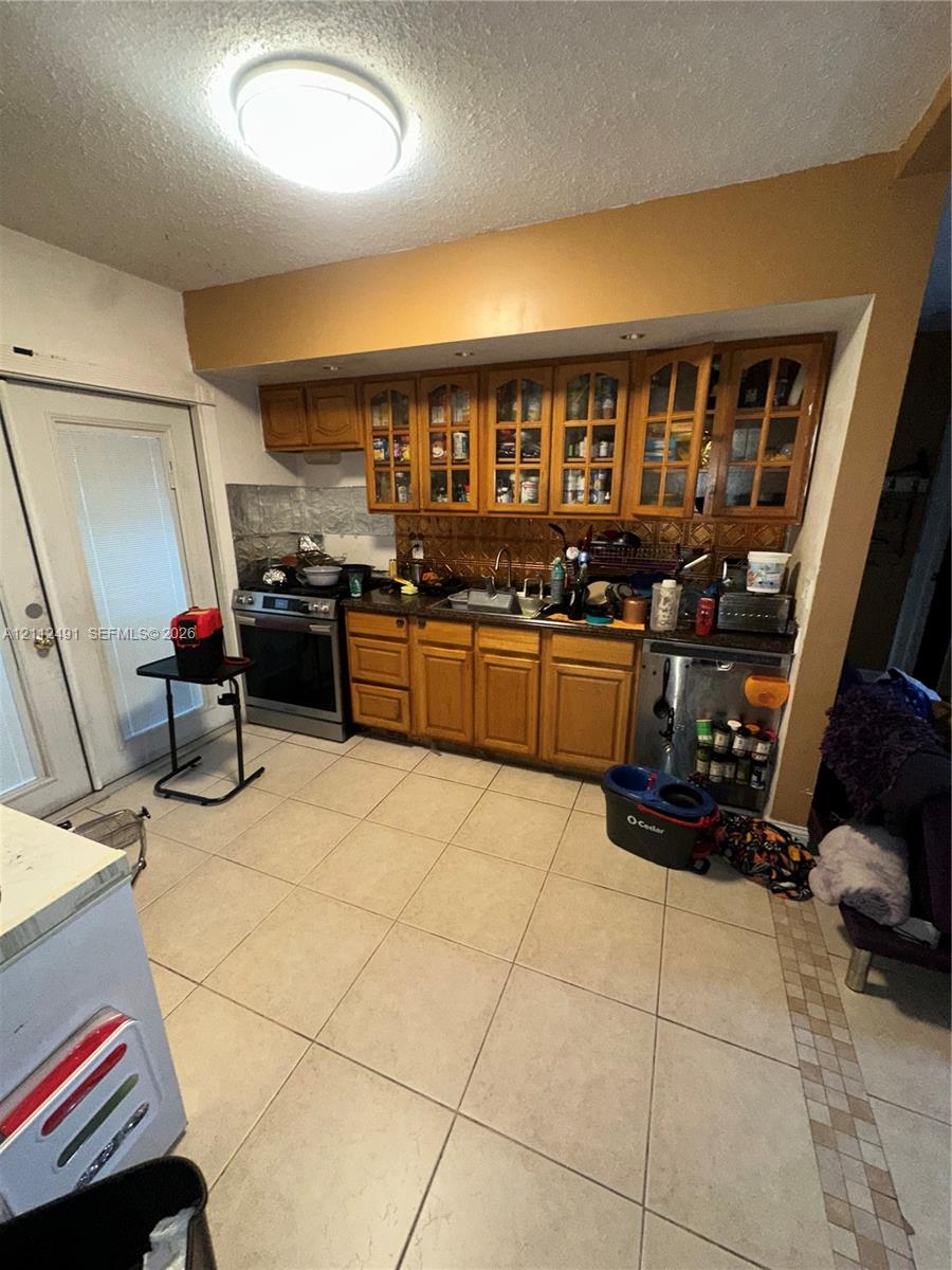 Kitchen featuring light wood cabinetry with glass-panel upper doors, stainless steel oven, stone-finish backsplash, and large-format ceramic tile flooring