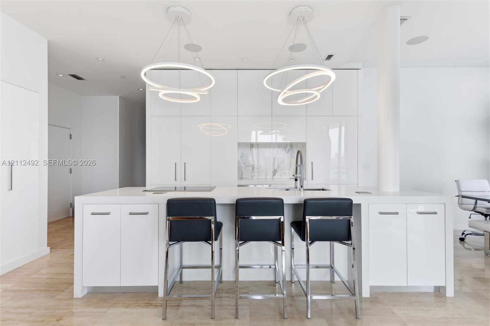 Modern kitchen featuring high-gloss cabinetry, a central island with a solid-surface countertop, dual contemporary ring pendant light fixtures, and stone tile flooring