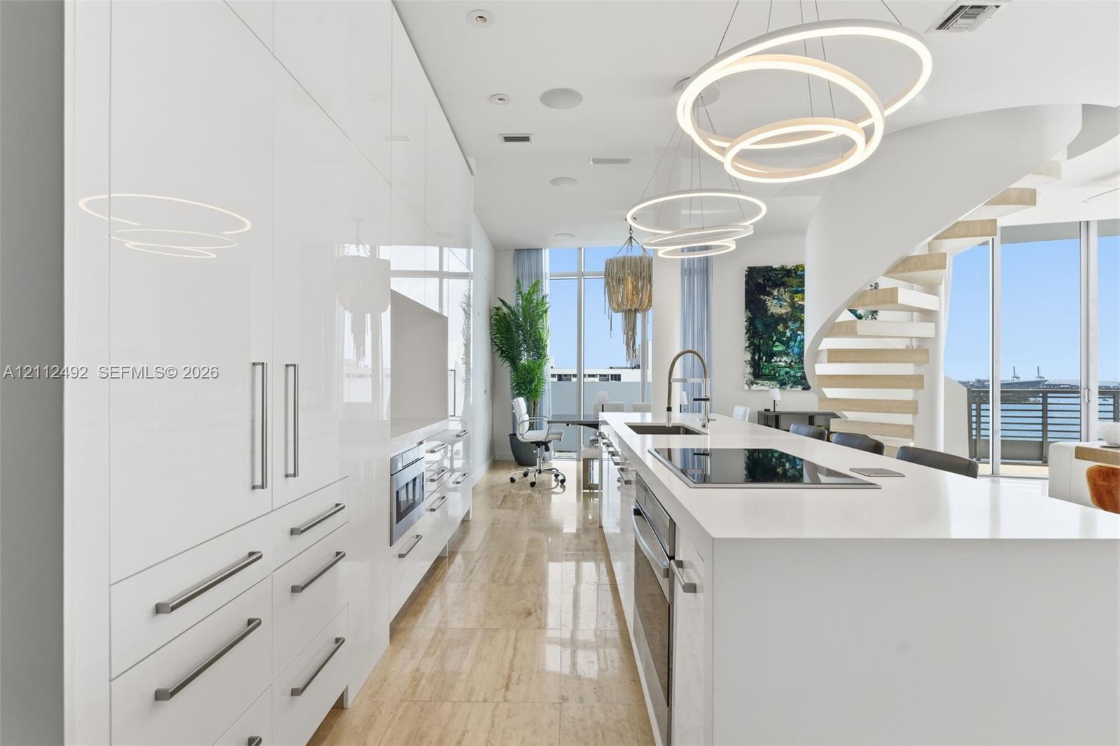 High-gloss cabinetry, stone-surfaced kitchen island, integrated stainless steel appliances, circular pendant lighting, and a sculptural spiral staircase with floating treads