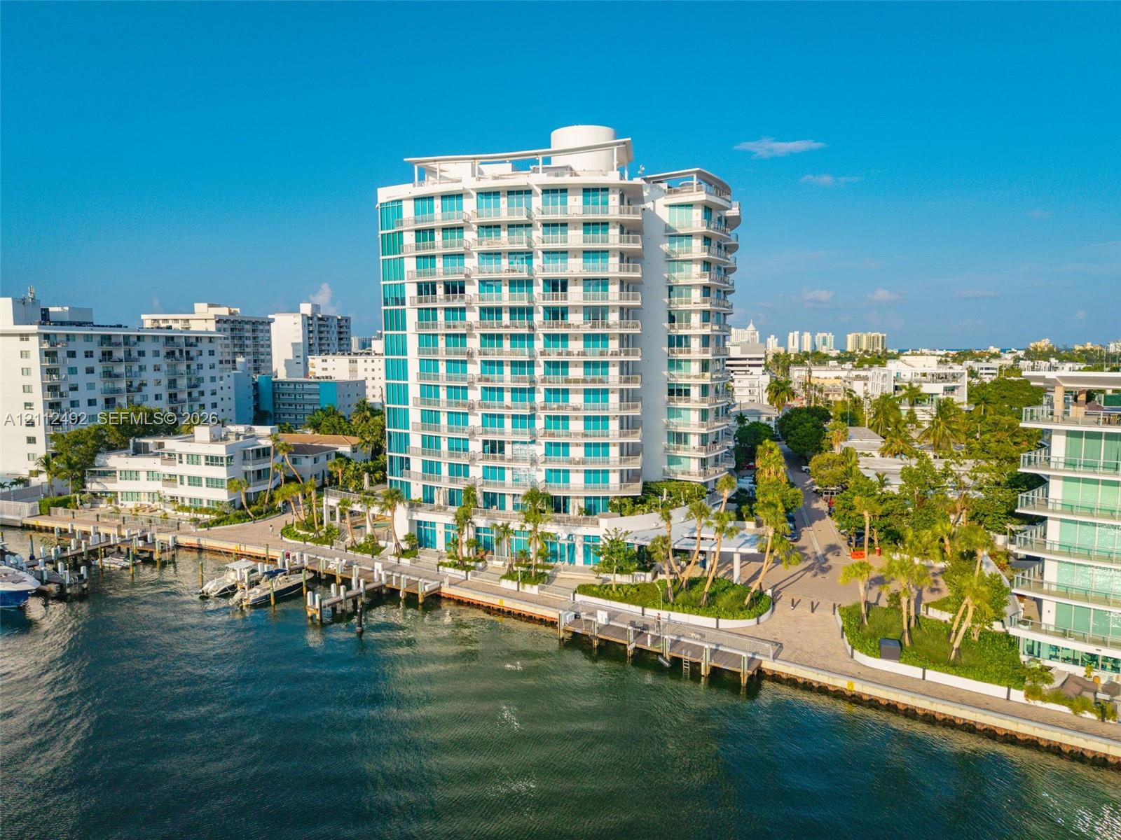 High-rise waterfront building featuring floor-to-ceiling glass paneling, tiered private balconies, an integrated deep-water dock, and paved perimeter hardscaping