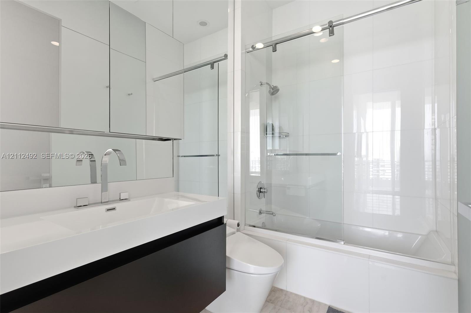 Modern vanity with integrated rectangular basin and polished chrome widespread faucet, mirrored medicine cabinet, floor-to-ceiling tiled shower enclosure with sliding glass door, and wall-mounted commode