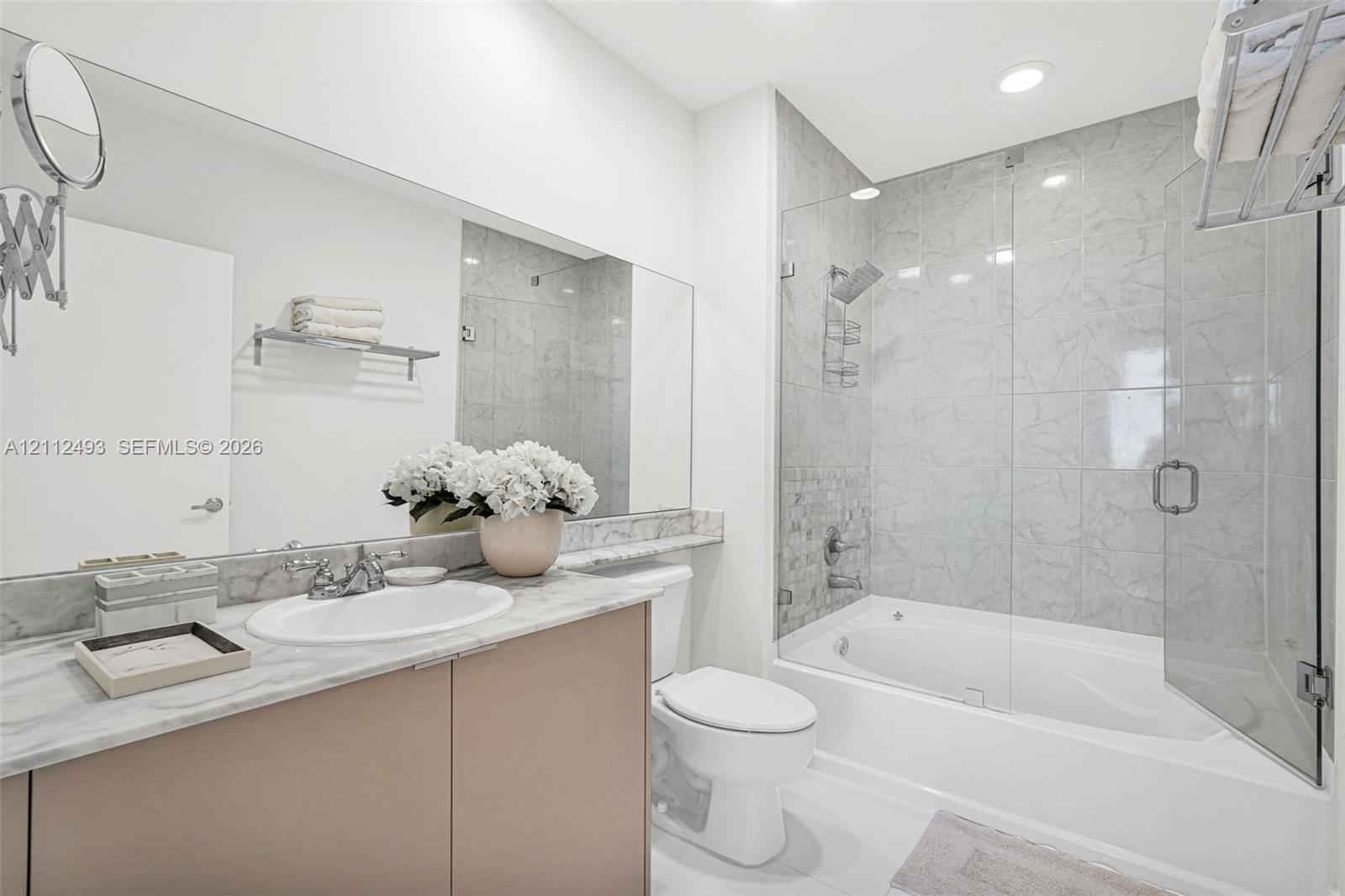 Bathroom featuring a vanity with a light stone countertop, an integrated sink, a large wall-mounted mirror, a tiled shower-tub combination with a glass enclosure, and recessed lighting