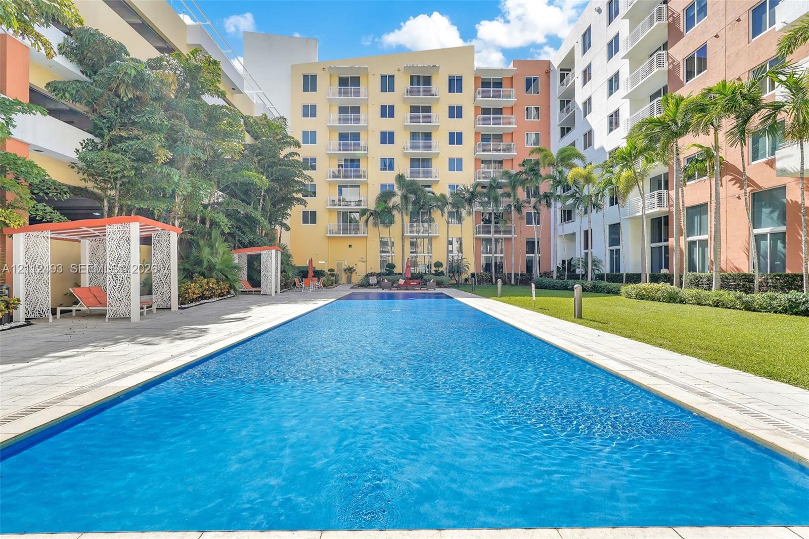 Rectangular swimming pool surrounded by stone paver decking, manicured lawn, architectural cabanas, and mature palm trees