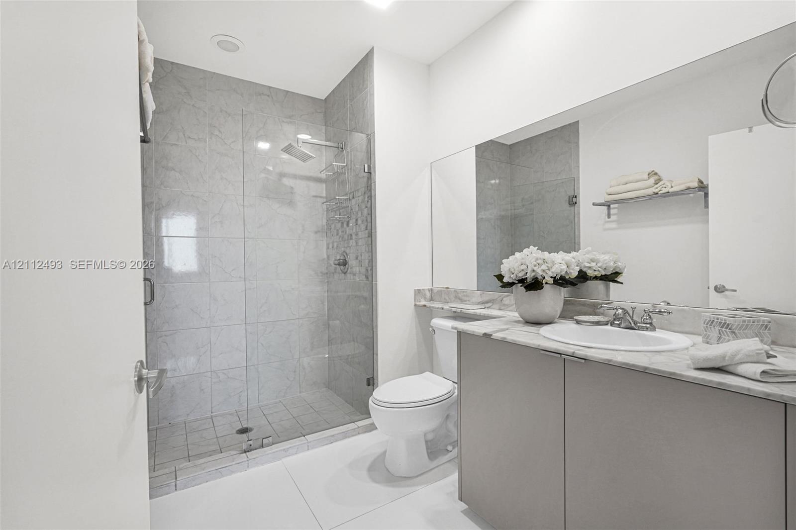 Walk-in shower with floor-to-ceiling tile, frameless glass enclosure, and rain-style showerhead, complemented by a vanity with stone-finish countertop and integrated sink