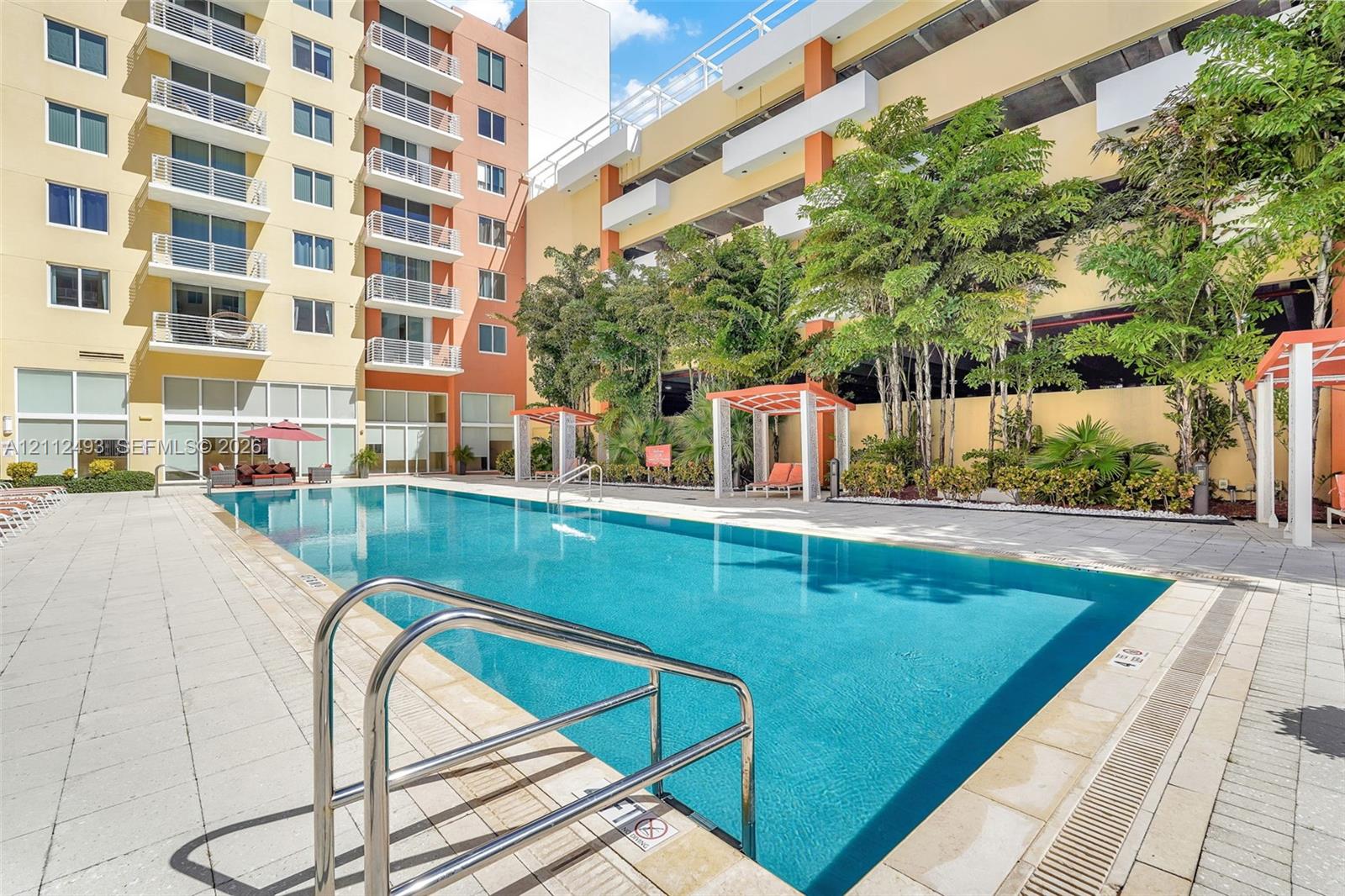 Swimming pool featuring a tiled deck, metal handrails, and structural cabana accents surrounded by palm trees